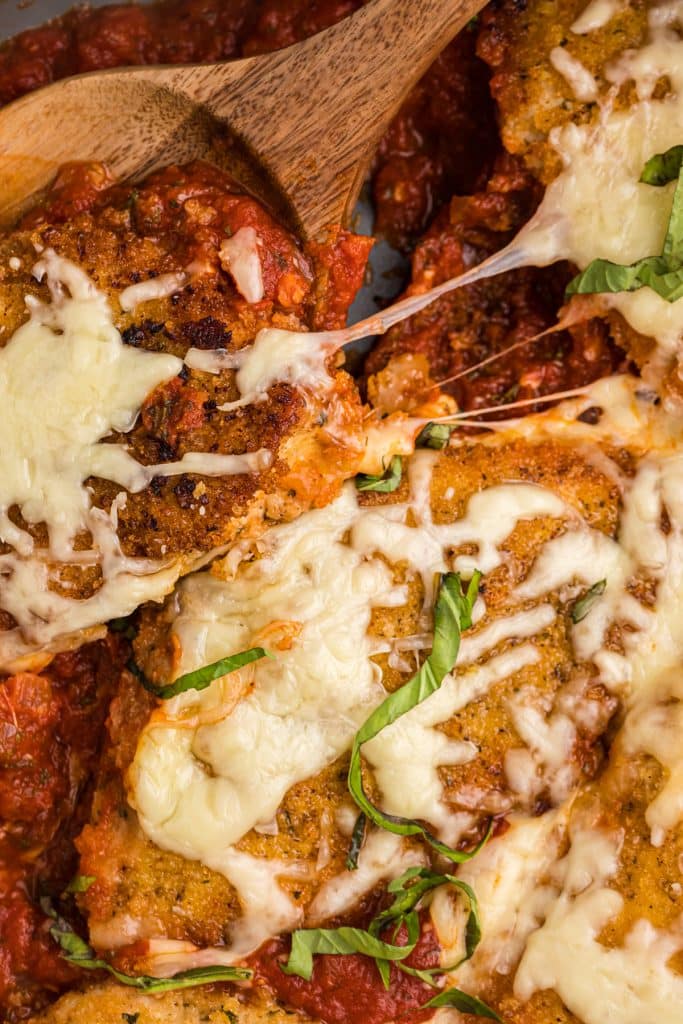 spoon lifting a chicken breast from a skillet with stretchy melted mozzarella cheese.