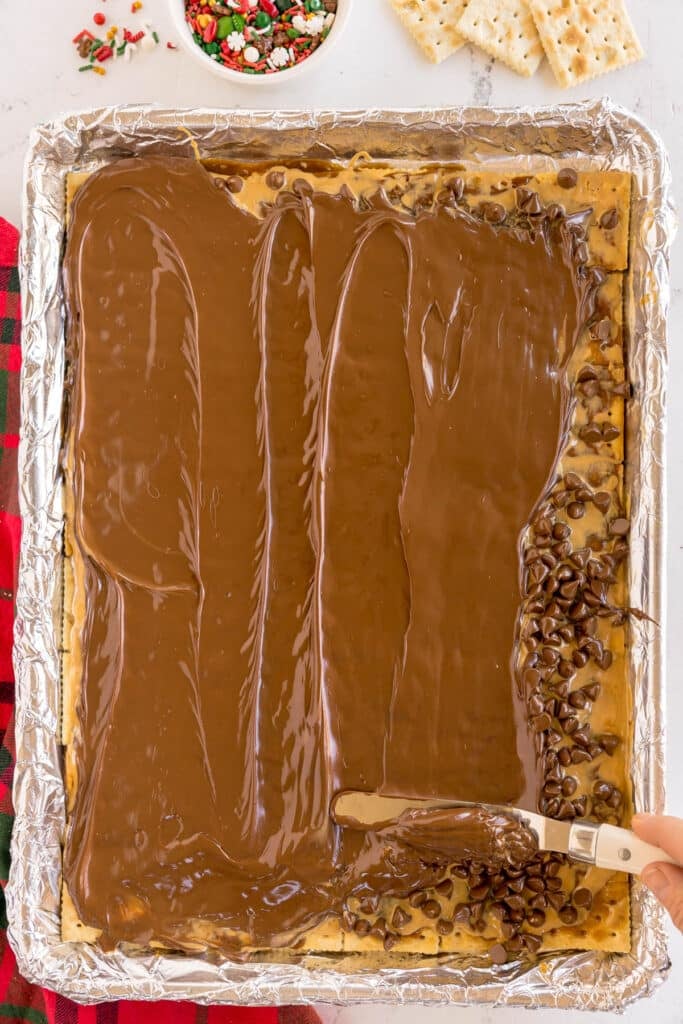 Melted chocolate being spread over a cracker layer in a baking tray.