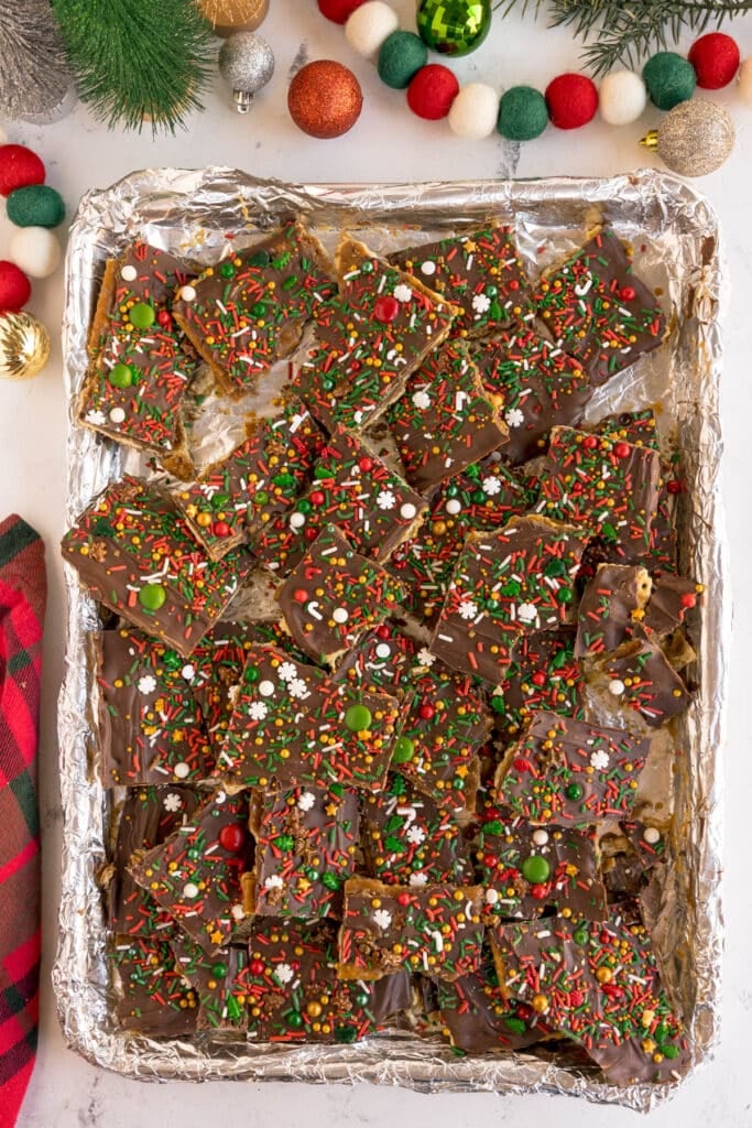 A baking tray of pieces of chocolate-covered Christmas crack decorated with colorful holiday sprinkles.