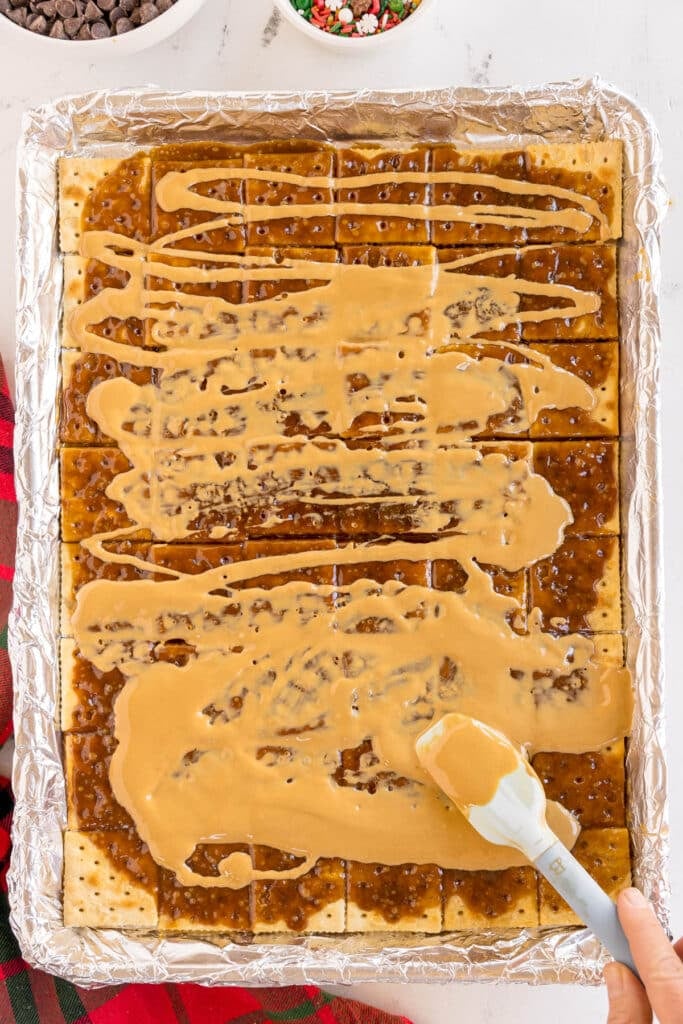 A baking tray with caramel-coated saltine crackers being drizzled and spread with smooth peanut butter, surrounded by ingredients like chocolate chips and holiday sprinkles.
