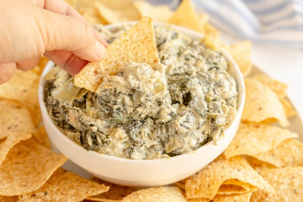 hand dipping tortilla chip into bowl of artichoke & spinach dip.