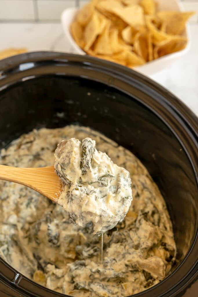 wooden spoon lifting spinach artichoke dip from a crockpot.