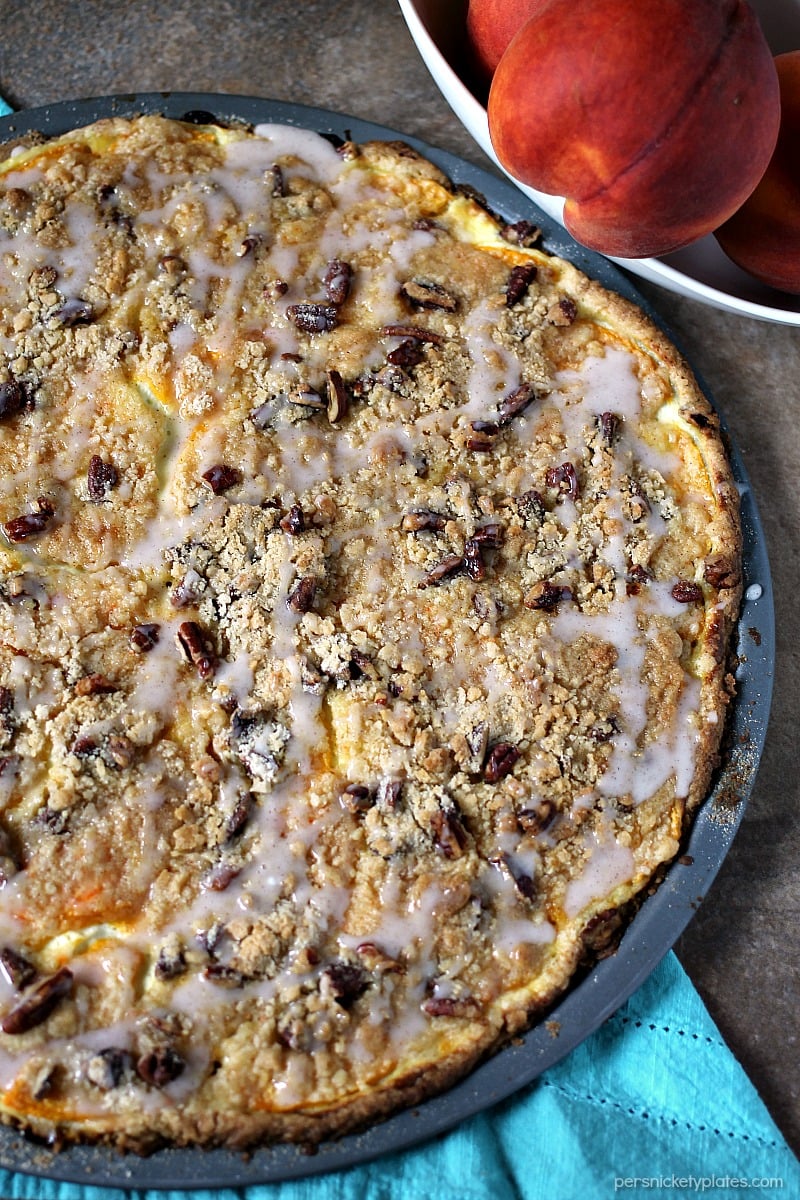 Peach Crumble Fruit Pizza is made on a cake batter crust, piled with thinly sliced fresh peaches and topped with a crumble and pecans and drizzled with a cinnamon sugar glaze. | www.persnicketyplates.com