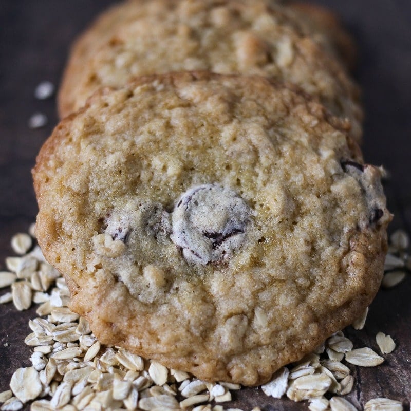 three ranger cookies on a dark wood table and spilled oats.
