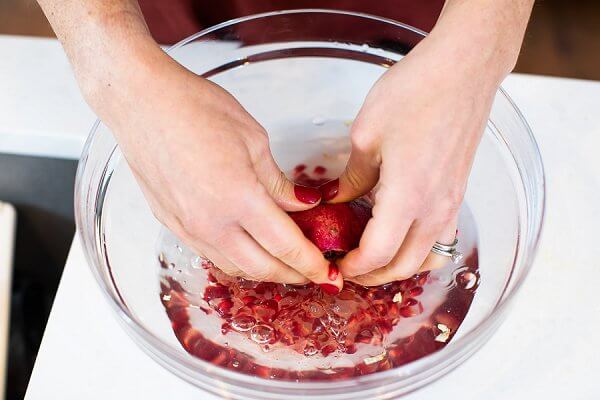 A large glass bowl half filled with water with half a pomegranate, and two hands removing the seeds from the pomegranate.