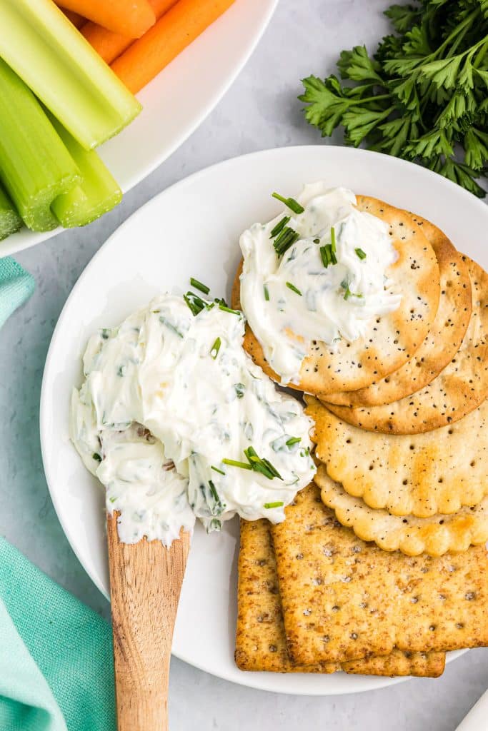 Garlic Herb DipGarlic Herb Dip starts with a cream cheese base then is filled with fresh herbs. Pair this super easy dip recipe with crackers, chips, or veggies to make the perfect quick snack for your next party or game day. | www.persnicketyplates.com #dip #appetizer #easyrecipe #partyfood #garlic knife spreading cream cheese dip on crackers.