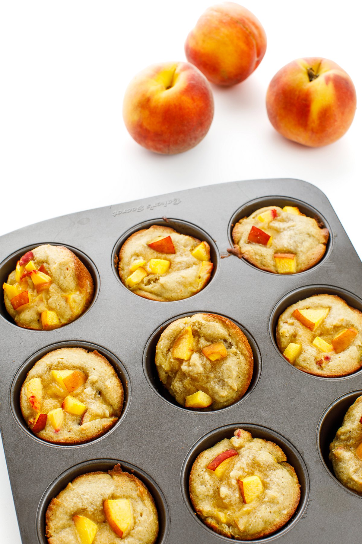 Almond flour peach muffins in a muffin tin next to three peaches.