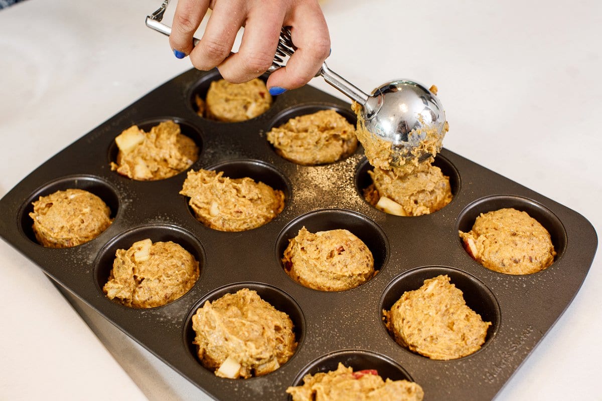 Liz's hand holding a scoop full of pumpkin muffin batter being poured into a muffin tin