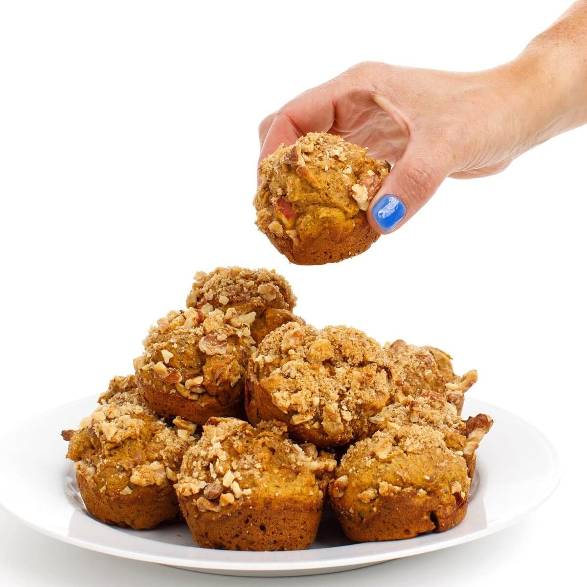 A hand grabbing a pumpkin applesauce muffin from a stack with a white background