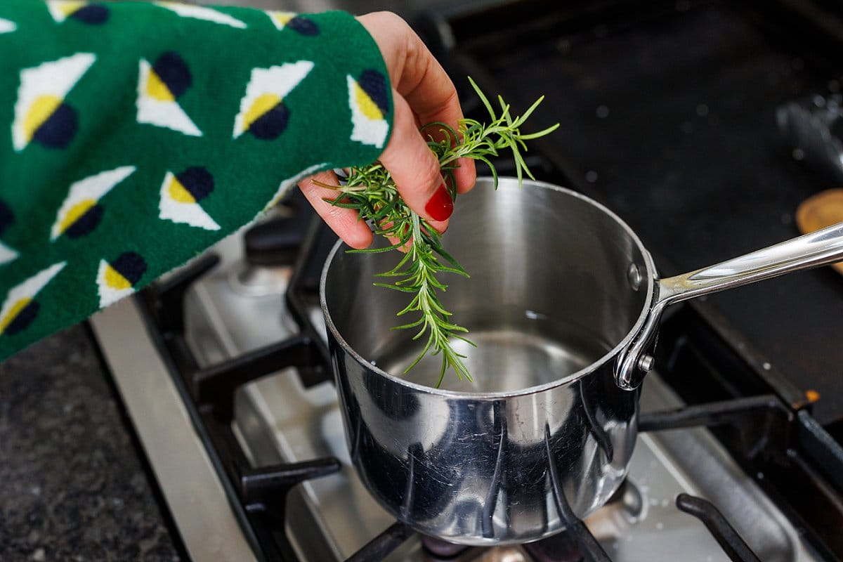 Adding rosemary sprigs to sugar and water in saucepan on the stove.