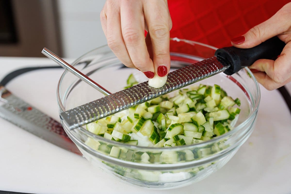 Using a microplane to grate garlic into a bowl with cucumber.