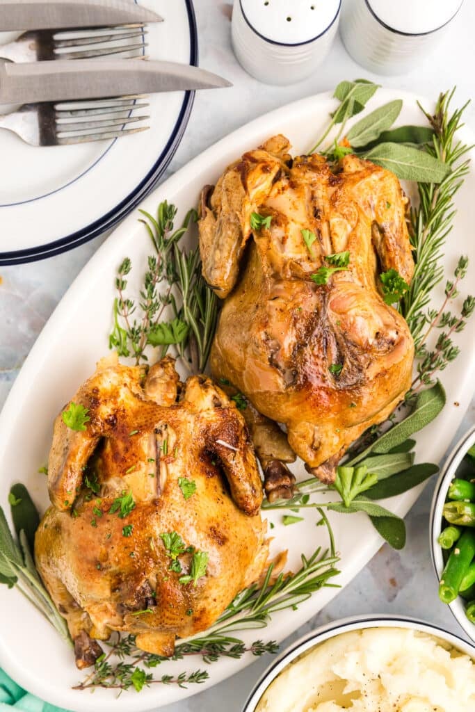 Two golden-brown Cornish hens placed on a white serving dish, surrounded by fresh rosemary, sage, and parsley, accompanied by green beans and mashed potatoes.