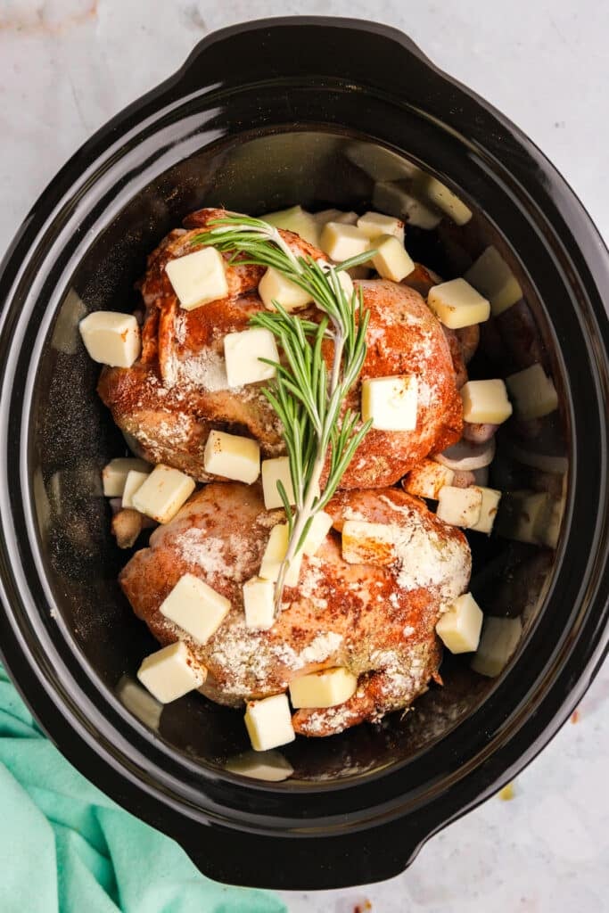 Two raw Cornish hens in a crockpot, seasoned and topped with small butter cubes and sprigs of fresh rosemary, ready to slow cook.