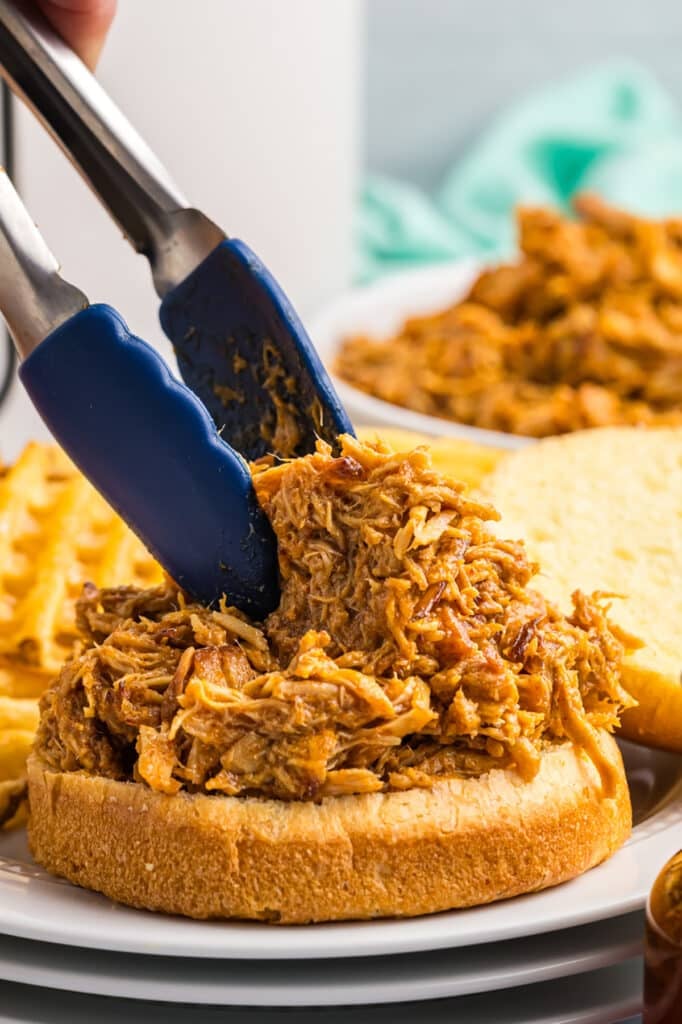 tongs piling shredded chicken onto a bun.