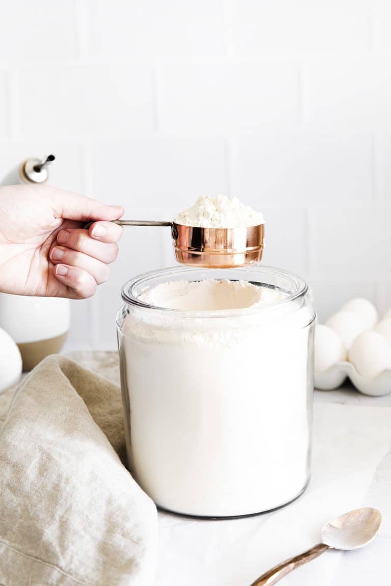 full measuring cup of flour being held over a glass jar of flour.