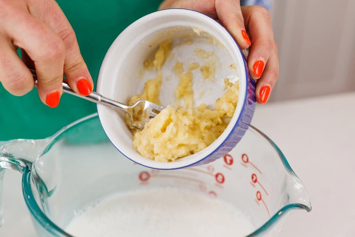 Spilling mashed banana out of a small glass dish into a large measuring bowl.