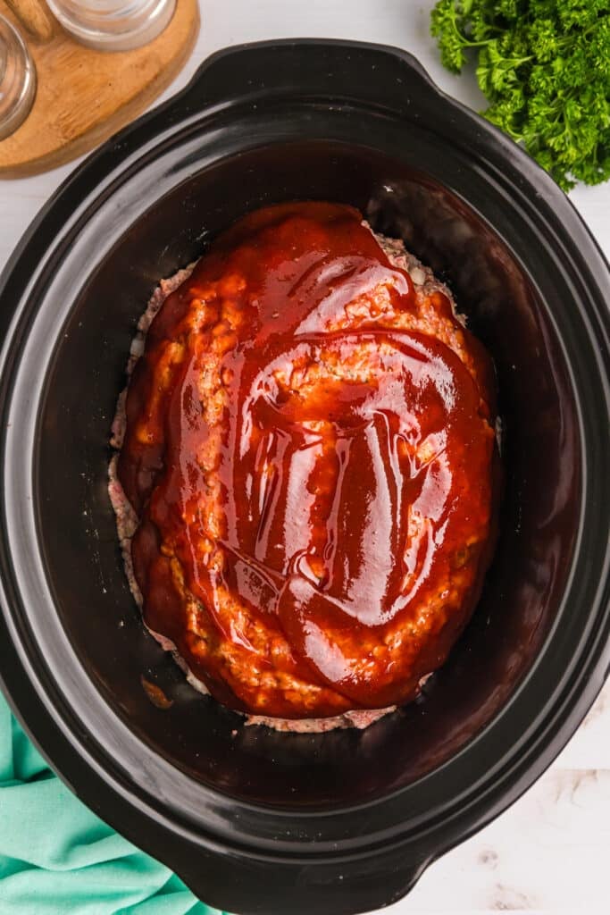overhead shot of sauce brushed onto a raw meatloaf in a slow cooker.