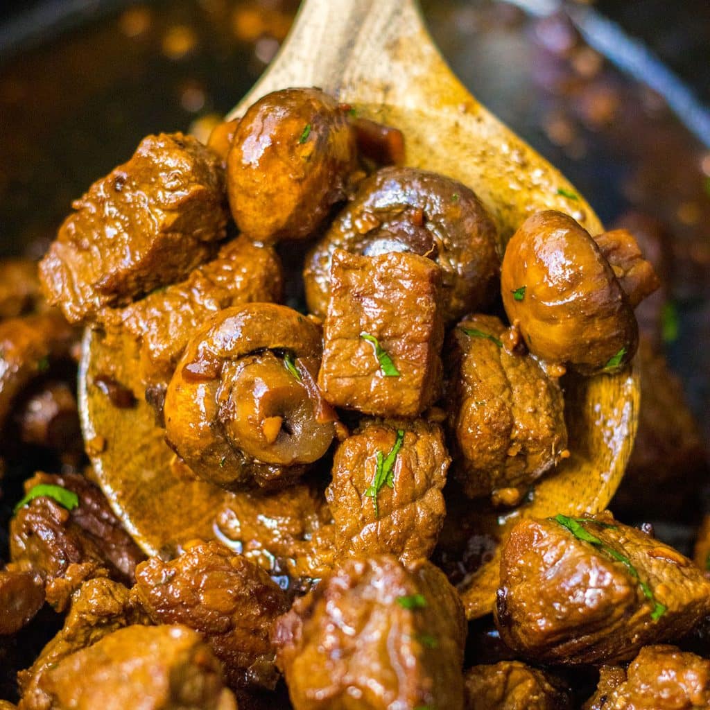 wooden spoon lifting a scoop of mushroom steak bites from a slow cooker.