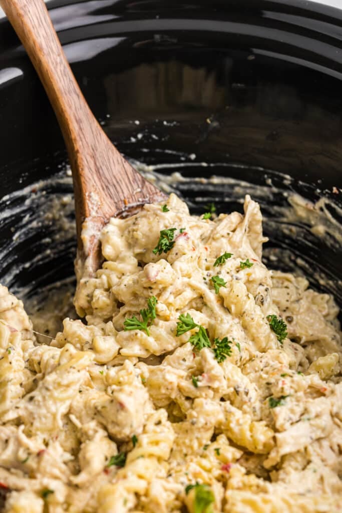 wooden spoon scooping chicken pasta from a slow cooker.