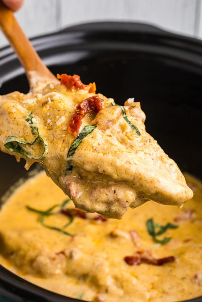 spoon lifting a chicken breast topped with tomatoes & basil from a slow cooker.