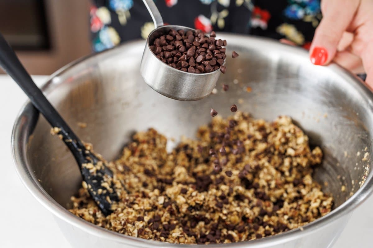 Adding chocolate chips to granola bar mixture in a large metal bowl.
