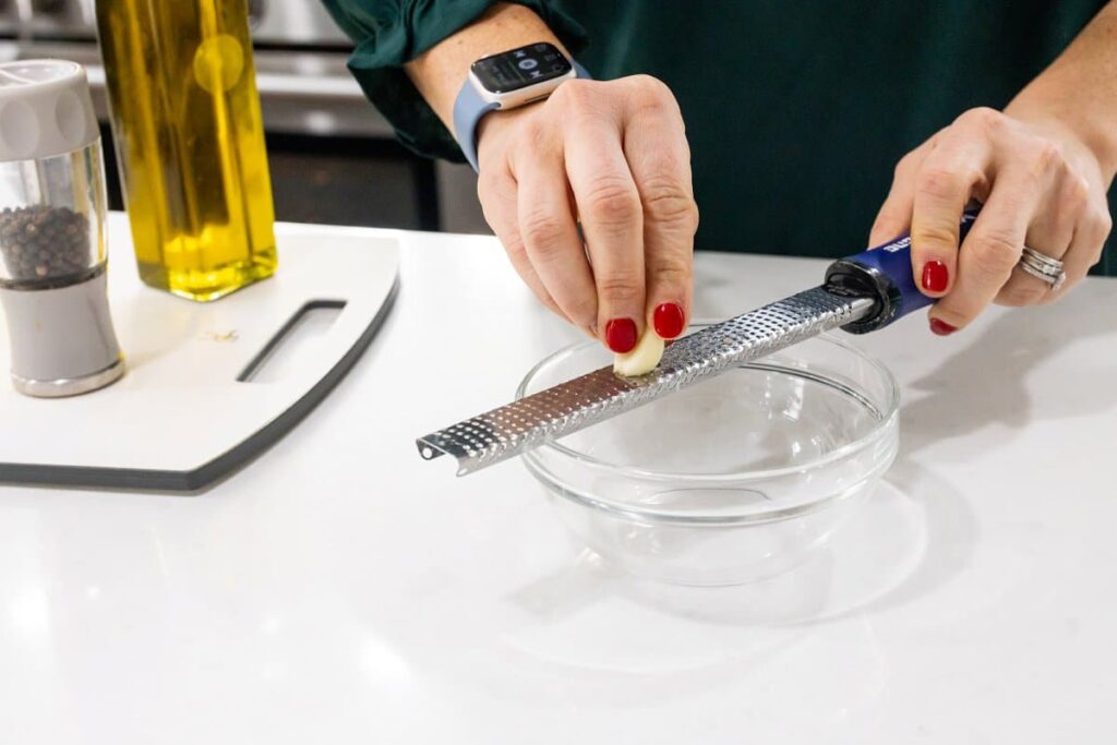 Grating a garlic clove into a small glass bowl with a microplane.
