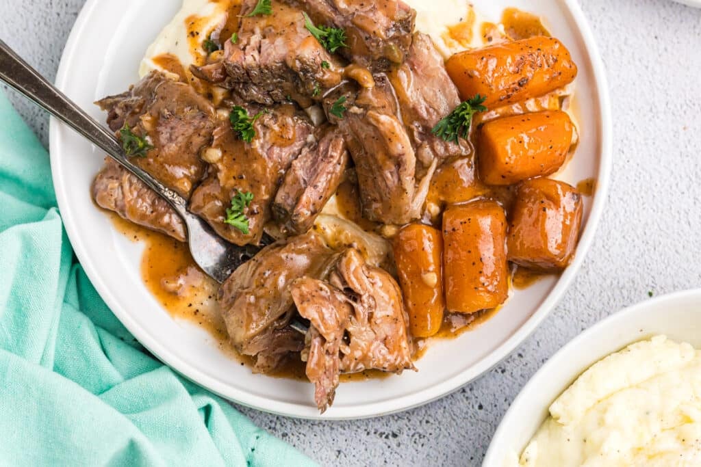 fork taking a bite of short rib, potatoes, and carrot on a plate.