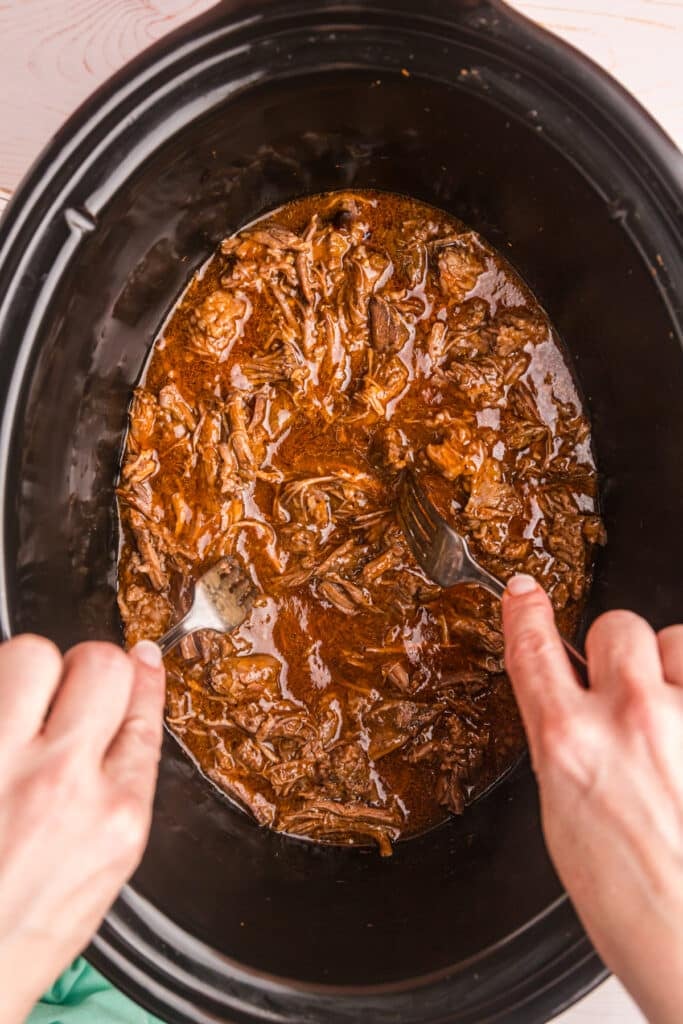 hands using forks to shred beef in a crockpot.