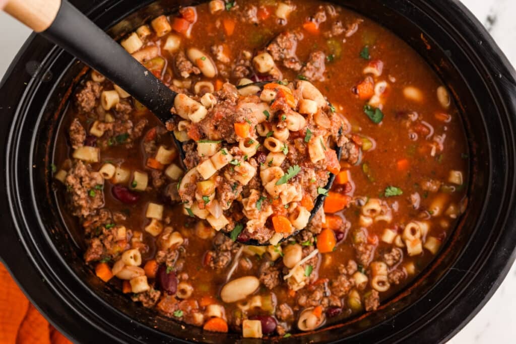 ladle lifting a scoop of pasta e fagioli soup from a crockpot.