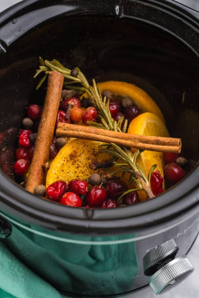 A black crockpot filled with sliced oranges, cranberries, cinnamon sticks, and spices.
