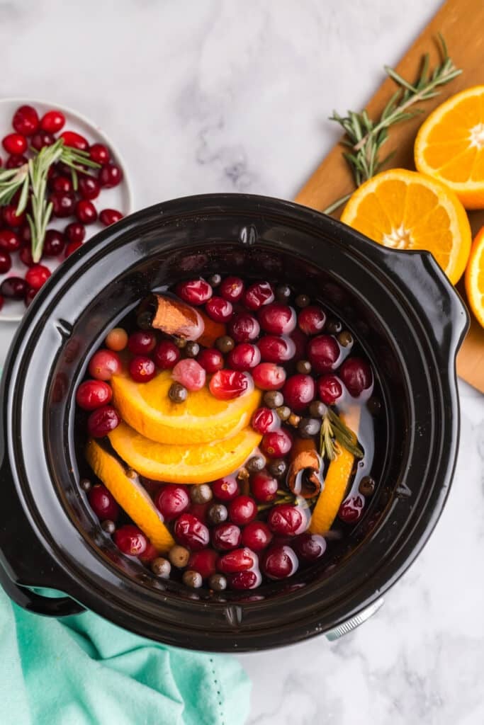 overhead shot of a black slow cooker filled with sliced oranges, cranberries, cinnamon sticks, and spices.