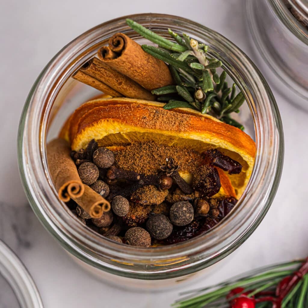 overhead shot of dried orange, cinnamon sticks, and spices in a glass jar.