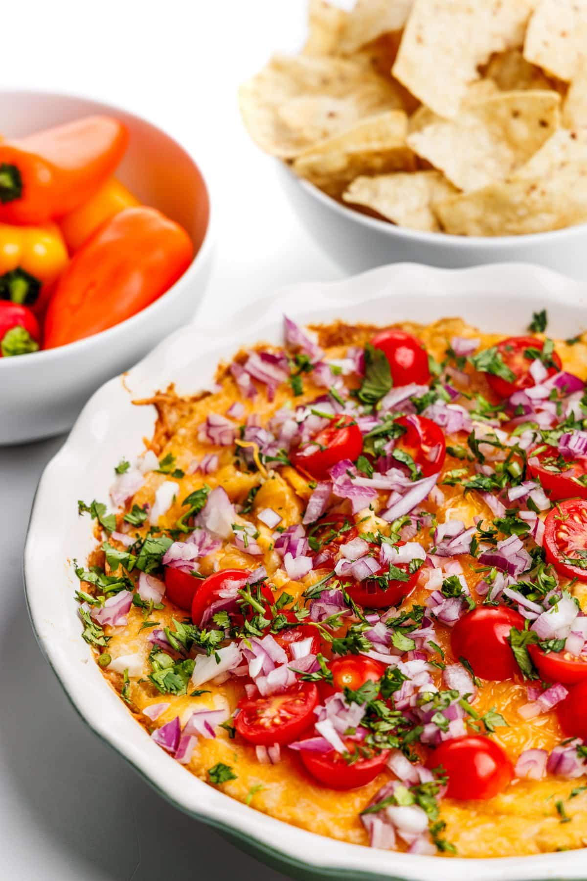 A close up view of protein packed baked hummus dip next to a bowl of tortilla chips and a bowl of mini peppers.