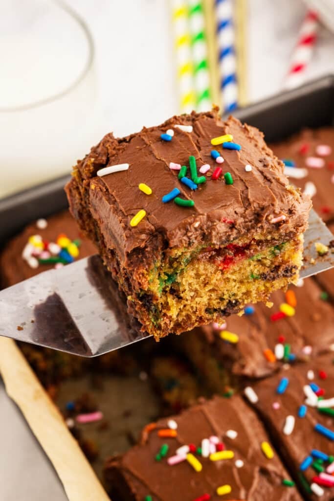 Slice of confetti cake with colorful sprinkles in the batter, topped with chocolate frosting and sprinkles, being held on a spatula above a pan.