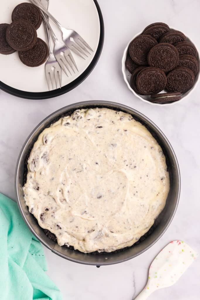 Overhead view of a cheesecake batter spread evenly in a pan, with visible chocolate cookie chunks mixed into the creamy filling.
