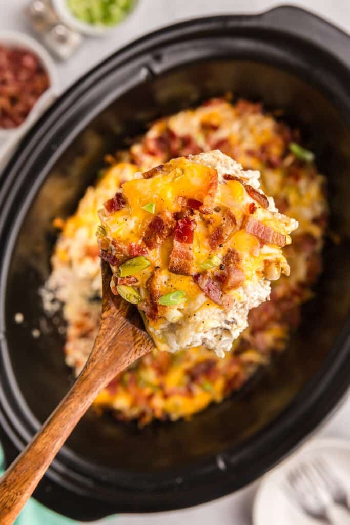 A wooden spoon lifting hash brown casserole from a crockpot.