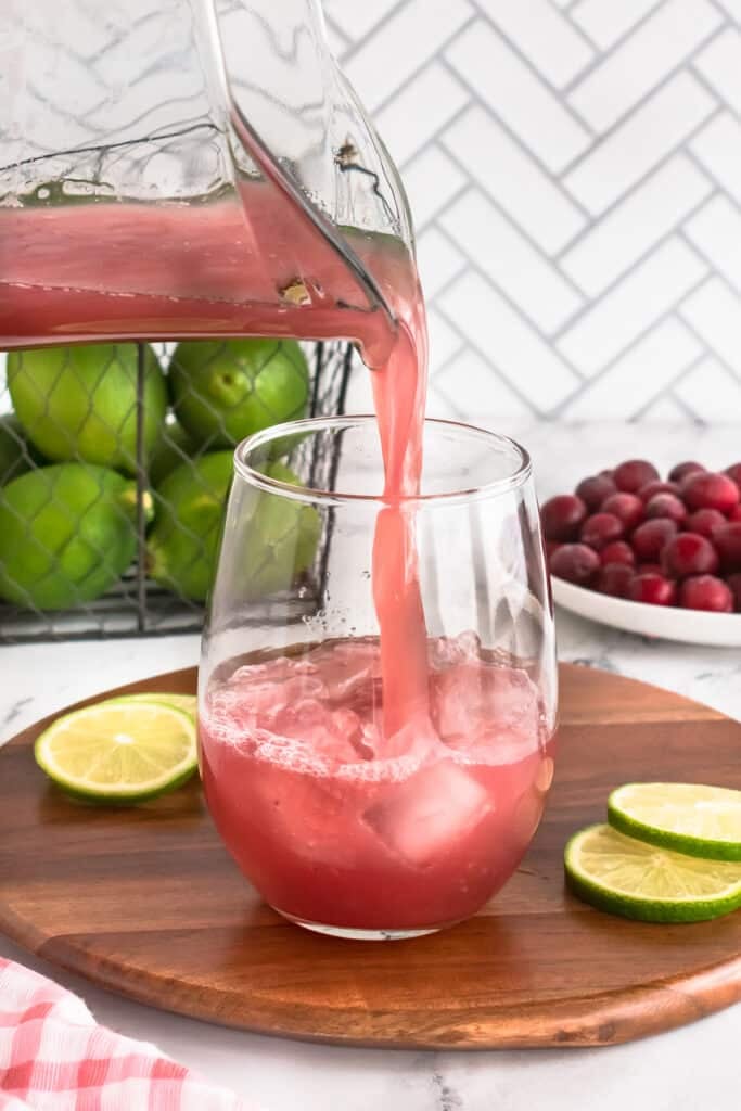 Cranberry punch being poured from a pitcher into a glass with ice, lime slices, and cranberries in the background.