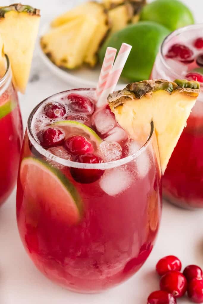 A glass of cranberry punch with lime slice, cranberries, pineapple garnish, and pink-striped straws on a wooden tray.