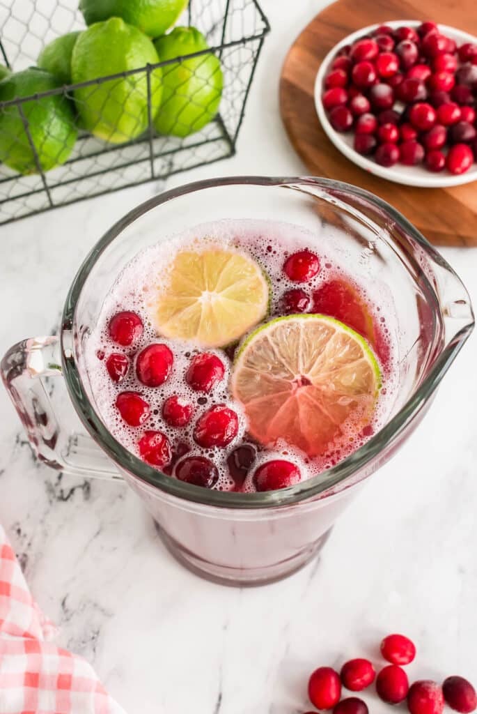 A pitcher of cranberry punch with lime slices and cranberries on top, surrounded by fresh limes and cranberries.