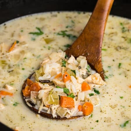 Close-up of a wooden ladle lifting a portion of creamy turkey and rice soup, showing chunks of turkey, diced carrots, celery, and parsley in a creamy broth.