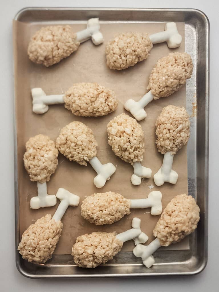 A tray of Rice Krispies treats shaped like turkey legs with white candy bones on parchment paper.
