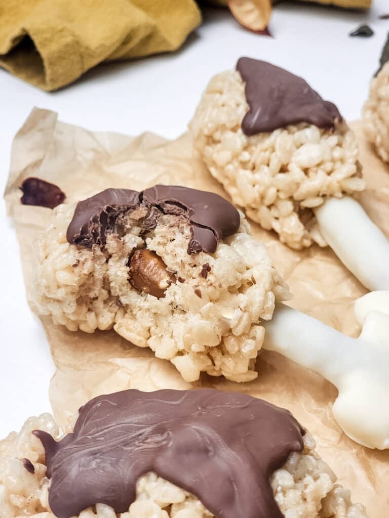 Close-up of Rice Krispies treats molded to look like turkey legs with chocolate drizzled on top, set on a crinkled parchment paper background. The "bone" part is white-coated and extends from the treat.
