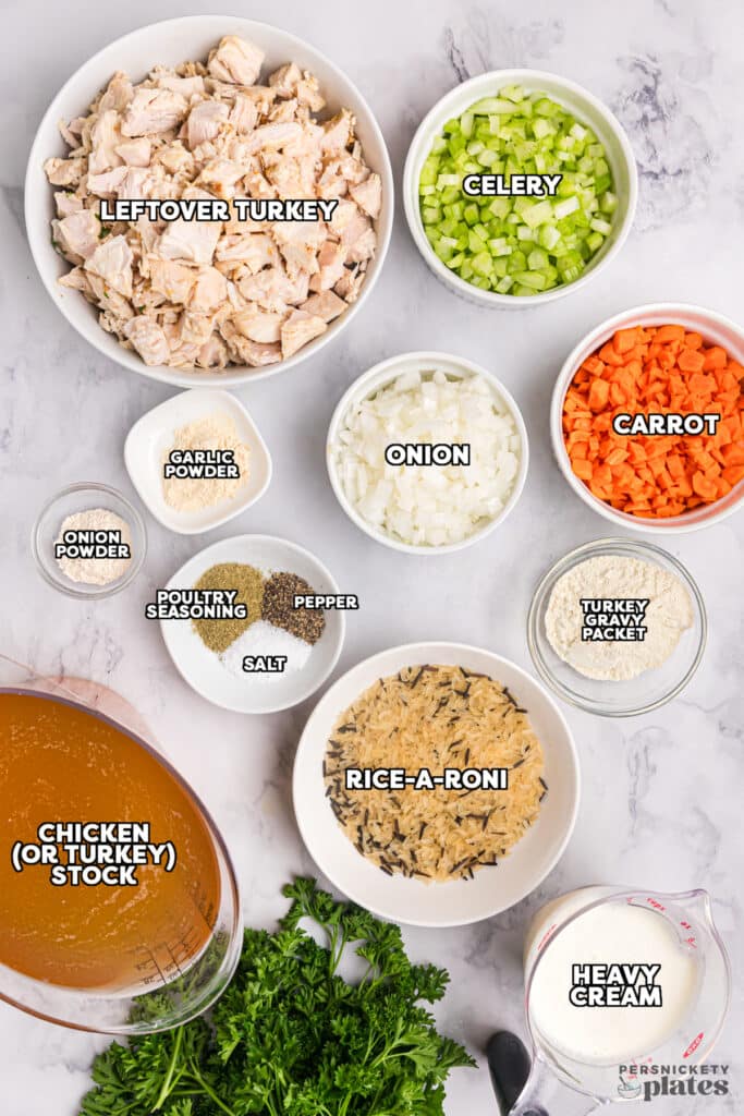 A variety of ingredients for turkey rice soup displayed in small bowls on a marble surface, including chopped leftover turkey, diced celery, carrots, and onions, along with seasonings, rice-a-roni, stock, and heavy cream. Labels identify each ingredient.