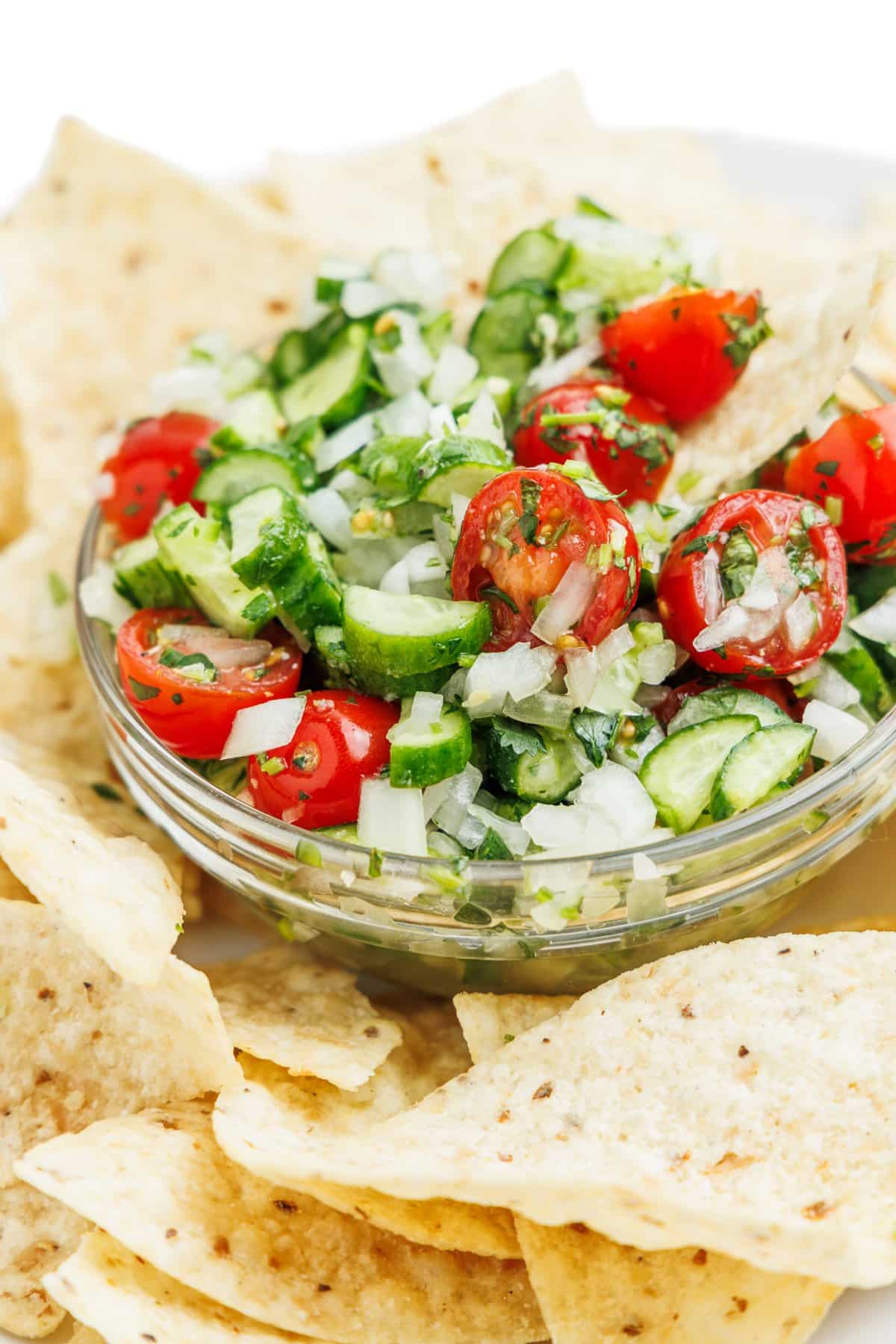A close up image of cucumber pico de gallo in a small glass bowl with a single tortilla chip in it.