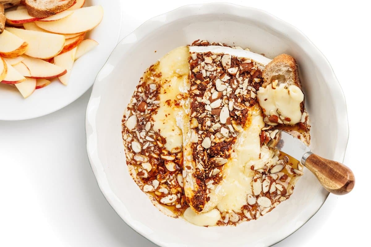 Hot honey baked brie in a baking dish with a knife and piece of bread, next to a plate with sliced apples.