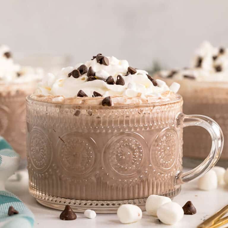 A glass mug filled with hot chocolate topped with whipped cream, mini chocolate chips, and marshmallows.