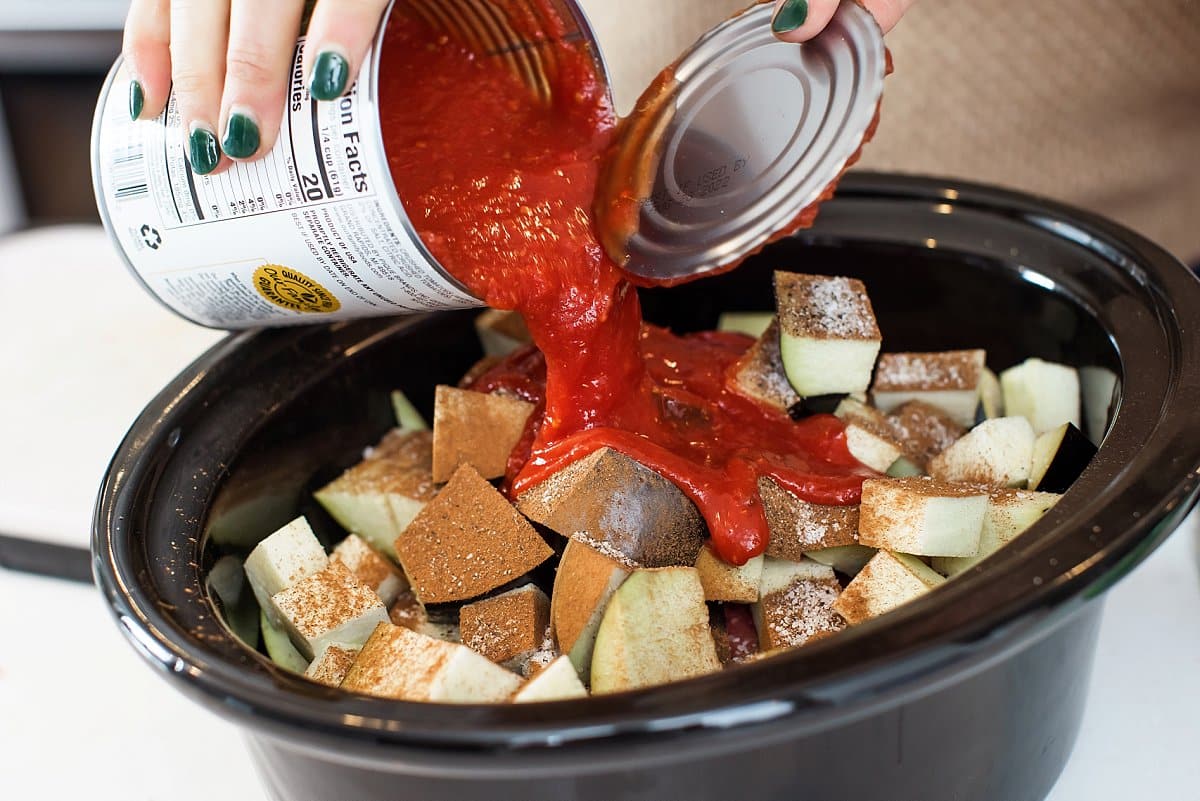 Adding crushed tomatoes to eggplant in slow cooker bowl.