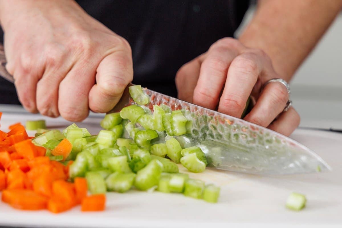 Liz dicing celery and carrot on a cutting board with a large, sharp knife.