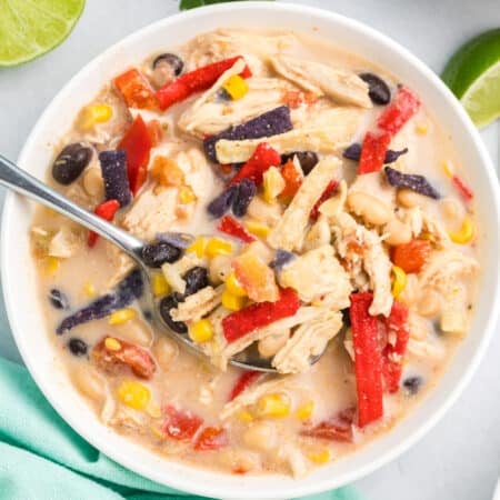 Close-up of a bowl of chicken tortilla soup with shredded chicken, beans, corn, and colorful tortilla strips, with fresh cilantro in the background.