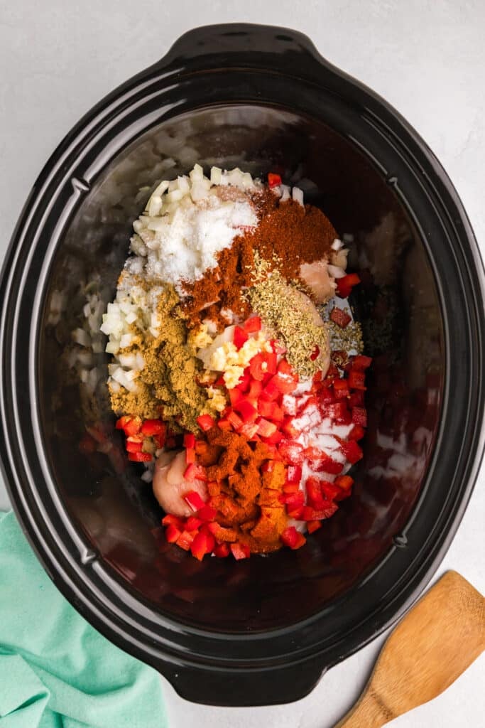 overhead shot of chicken breasts, diced peppers, onions and seasonings, in a black crockpot.